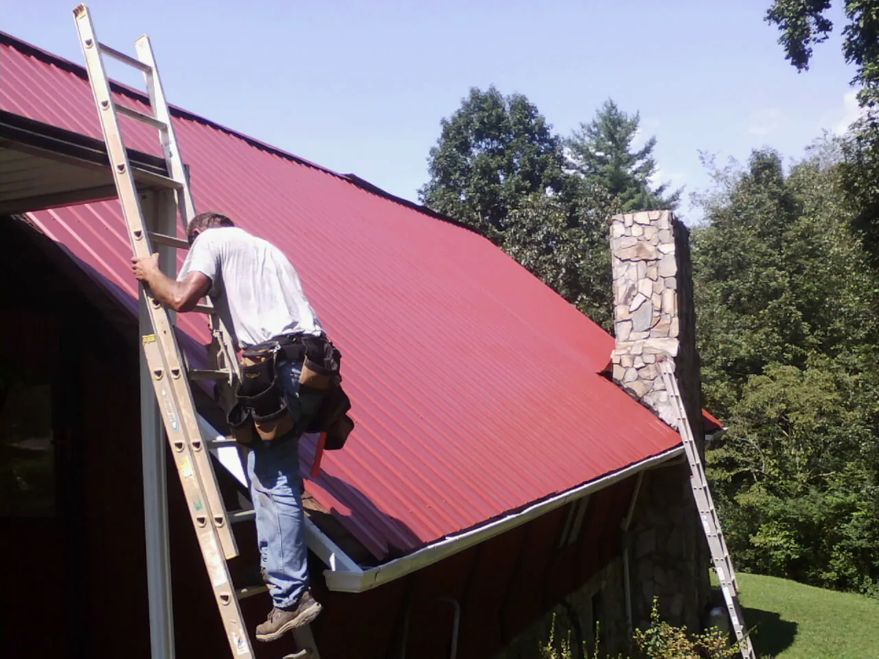 Roofer on a ladder beside a red metal roof and stone chimney — Soffit & Fascia Repair by Roofing Brothers in Blackstock